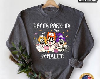 May include: Dark gray heather sweatshirt with the text "Hocus Poke-Us" and the hashtag "#CNALIFE." The design features three cartoon ghosts dressed as nurses, holding medical items. The sweatshirt has a crew neck and long sleeves.