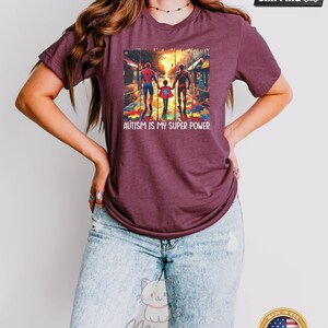 May include: A heather maroon t-shirt with a graphic of three figures walking on a street with a puzzle piece pattern on the ground. The text "AUTISM IS MY SUPER POWER" is printed below the graphic.
