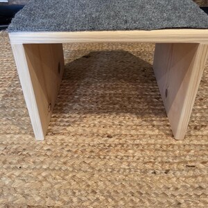 May include: A low wooden stool with a rectangular top covered in a textured gray felt fabric. The base is constructed from light-colored natural wood with visible grain patterns, featuring two side panels that support the flat, elevated surface.