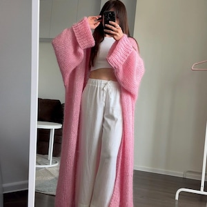 Pode incluir: Cardigã comprido de malha rosa oversized com mangas largas. O cardigã é combinado com calças brancas e um top branco. A roupa é refletida num espelho.