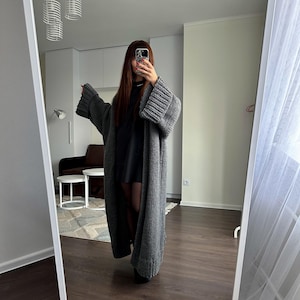 May include: A long, gray, open-front cardigan with wide sleeves. The chunky knit sweater is floor-length and appears to be made of a soft, warm material. The cardigan is being worn in a full-length mirror.