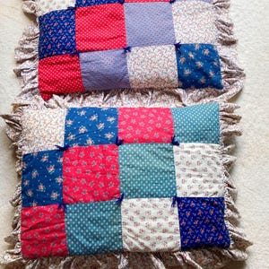Puede incluir: Dos cojines rectangulares de patchwork con bordes con volantes. Cada cojín presenta una cuadrícula de cuadrados coloridos con patrones florales rojos, azules y blancos. Los cuadrados están conectados con pequeños lazos azules. Los bordes con volantes tienen un estampado floral.