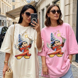 Peut inclure: Deux femmes portant des t-shirts amples avec un motif de Mickey Mouse magicien. Un t-shirt est cr&egrave;me, l'autre rose. Les deux t-shirts pr&eacute;sentent un Mickey Mouse de dessin anim&eacute; avec une baguette. Les femmes portent des lunettes de soleil et des pantalons ray&eacute;s.