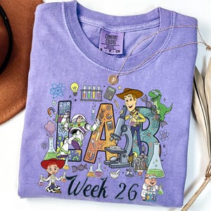 Op de afbeelding: Een lavendelkleurig Comfort Colors shirt met lange mouwen met een kleurrijke afbeelding van Toy Story-personages in een wetenschappelijk laboratorium. Het ontwerp bevat de tekst "LAB" en "Week 26". Een gouden ketting en een bruine hoed staan ook op de afbeelding.