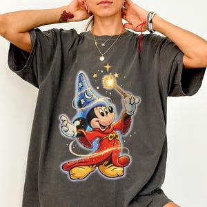 Peut inclure: T-shirt gris fonc&eacute; avec un motif color&eacute; de Mickey Mouse en magicien, tenant une baguette avec une &eacute;toile brillante. Le personnage de dessin anim&eacute; porte un chapeau de magicien bleu et une robe rouge. Le t-shirt a des manches courtes et une coupe d&eacute;contract&eacute;e.