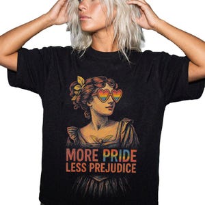 May include: Black t-shirt featuring a vintage illustration of a woman wearing heart-shaped sunglasses with rainbow lenses. The graphic includes the text "MORE PRIDE LESS PREJUDICE" in orange and blue. The design is a statement of LGBTQ+ pride.