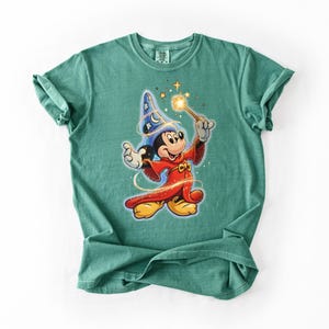 Peut inclure: T-shirt de couleur vert d'eau avec une illustration de Mickey Mouse en magicien. Mickey porte un chapeau de magicien bleu avec des &eacute;toiles et une robe rouge, tenant une baguette lumineuse. Le motif est centr&eacute; sur le t-shirt.