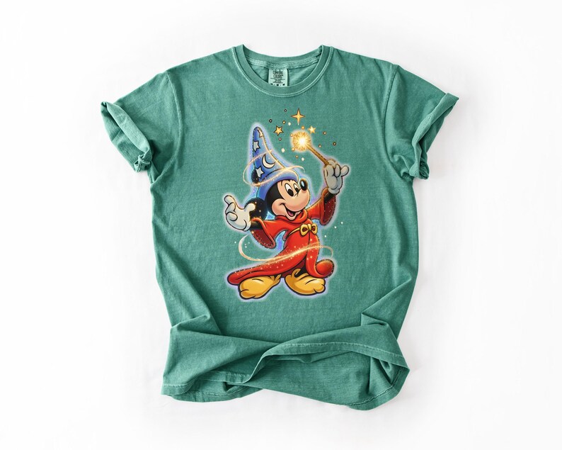 Peut inclure: T-shirt de couleur vert d'eau avec une illustration de Mickey Mouse en magicien. Mickey porte un chapeau de magicien bleu avec des &eacute;toiles et une robe rouge, tenant une baguette lumineuse. Le motif est centr&eacute; sur le t-shirt.