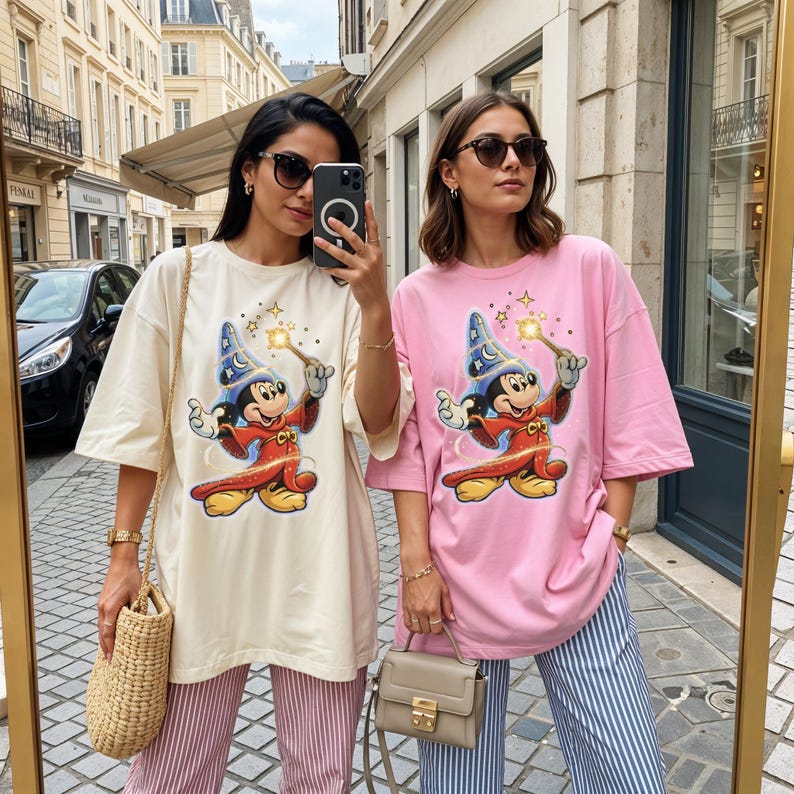Peut inclure: Deux femmes portant des t-shirts amples avec un motif de Mickey Mouse magicien. Un t-shirt est cr&egrave;me, l'autre rose. Les deux t-shirts pr&eacute;sentent un Mickey Mouse de dessin anim&eacute; avec une baguette. Les femmes portent des lunettes de soleil et des pantalons ray&eacute;s.