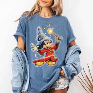 Peut inclure: T-shirt bleu avec un dessin de Mickey Mouse en magicien, tenant une baguette lumineuse. Mickey porte une robe rouge et un chapeau de magicien bleu avec des &eacute;toiles et des lunes. Le t-shirt a des manches retrouss&eacute;es.