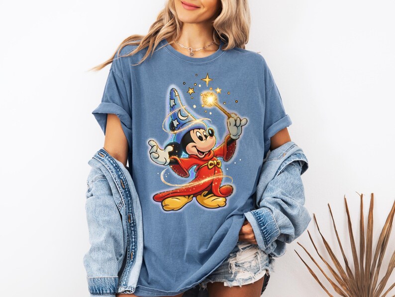 Peut inclure: T-shirt bleu avec un dessin de Mickey Mouse en magicien, tenant une baguette lumineuse. Mickey porte une robe rouge et un chapeau de magicien bleu avec des &eacute;toiles et des lunes. Le t-shirt a des manches retrouss&eacute;es.