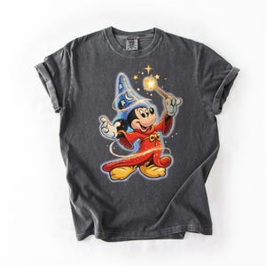 Peut inclure: T-shirt gris fonc&eacute; avec un dessin de Mickey Mouse en magicien. Mickey porte un chapeau de magicien bleu avec des &eacute;toiles, une robe rouge et tient une baguette lumineuse. Le t-shirt est de style unisexe.