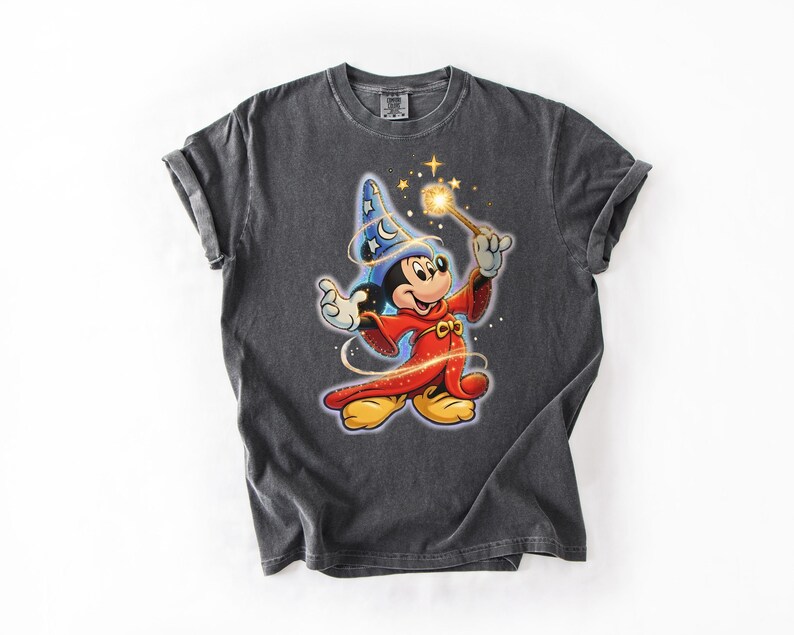Peut inclure: T-shirt gris fonc&eacute; avec un dessin de Mickey Mouse en magicien. Mickey porte un chapeau de magicien bleu avec des &eacute;toiles, une robe rouge et tient une baguette lumineuse. Le t-shirt est de style unisexe.