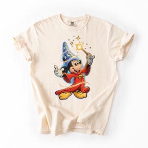 Peut inclure: T-shirt cr&egrave;me avec un dessin de Mickey Mouse en sorcier. Mickey porte un chapeau bleu &eacute;toil&eacute;, une robe rouge et tient une baguette magique dor&eacute;e. Le t-shirt a des manches retrouss&eacute;es.