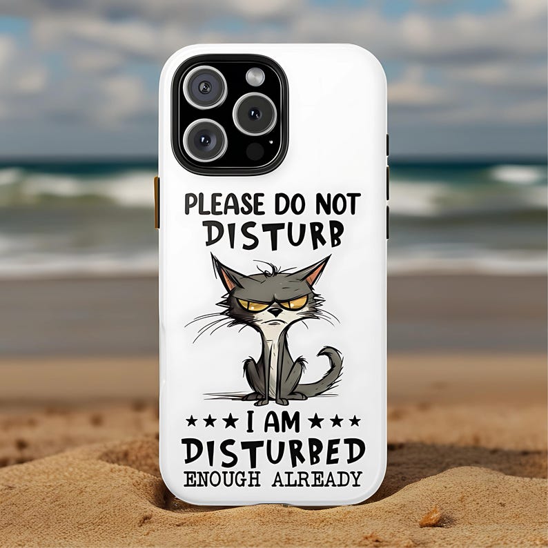 May include: White phone case with a black and gray cartoon cat illustration. The case has the text "PLEASE DO NOT DISTURB" above the cat and "I AM DISTURBED ENOUGH ALREADY" below it. The phone case is on a sandy beach.
