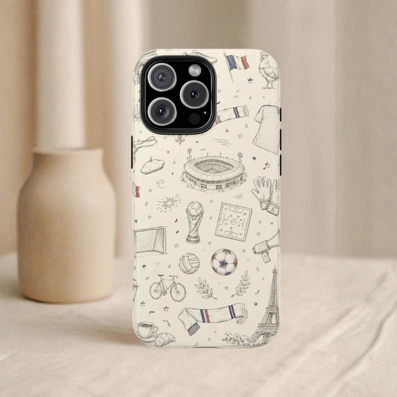 May include: A phone case with a cream background featuring a repeating pattern of French-themed illustrations. The drawings include a soccer ball, a stadium, a trophy, a bicycle, a croissant, and the Eiffel Tower.