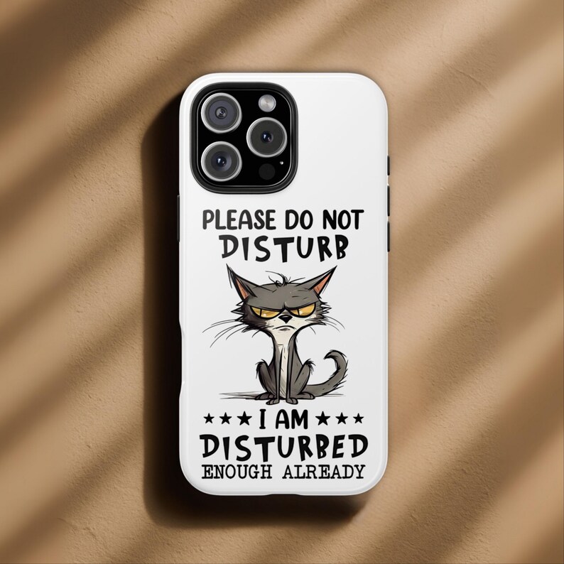 May include: White phone case with a cartoon illustration of an angry gray cat. The case has the text "PLEASE DO NOT DISTURB" above the cat and "I AM DISTURBED ENOUGH ALREADY" below it. The case has a glossy finish.