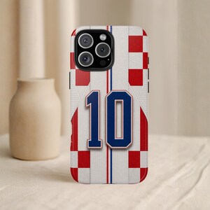 May include: A phone case featuring a design inspired by a soccer jersey. The case has a white background with red and blue accents, including the number 10 in blue. The design incorporates a checkered pattern.