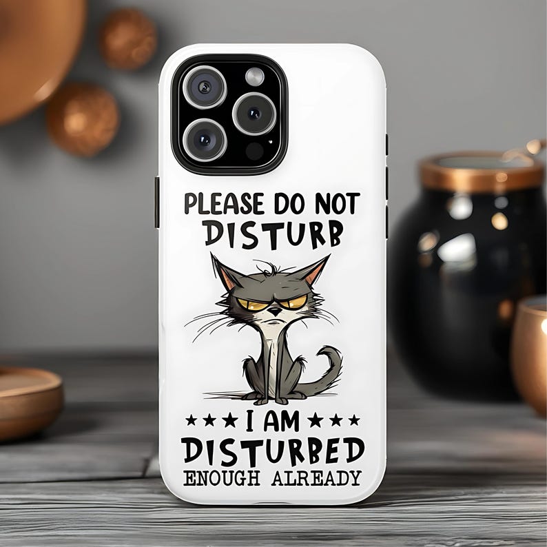 May include: White phone case with a black camera module and a cartoon cat illustration. The text on the case reads "PLEASE DO NOT DISTURB" above the cat and "I AM DISTURBED ENOUGH ALREADY" below.