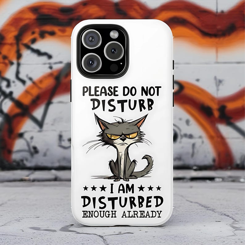 May include: White phone case with a cartoon image of a grumpy gray cat. The text on the case reads "PLEASE DO NOT DISTURB" above the cat and "I AM DISTURBED ENOUGH ALREADY" below it. The case has a black camera lens.