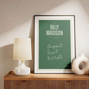 May include: A framed green poster with white text that reads "Billy Madison" and "dugout, bunt, pirruto". The poster is on a wooden shelf with a white lamp and a white ceramic vase.