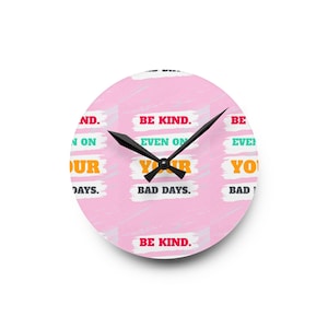 Op de afbeelding: Ronde wandklok met een roze achtergrond en de woorden "BE KIND" en "EVEN ON YOUR BAD DAYS" in verschillende kleuren. De klok heeft zwarte wijzers en een witte rand.