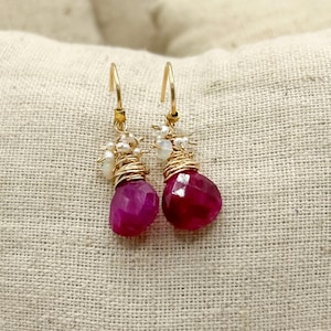 May include: A pair of gold-colored hoop earrings with dangling, faceted, magenta-colored gemstones. The gemstones are wire-wrapped with gold-colored wire and accented with small, white, pearl-like beads. The earrings are displayed against a neutral-toned background.