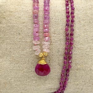 Könnte beinhalten: Eine Halskette mit einer Reihe facettierter rosa und hellrosa Perlen, die in einen größeren, tropfenförmigen, dunkelmagentafarbenen Edelsteinanhänger mit Golddrahtwicklung übergeht. Ein separater, längerer Strang aus kleinen, facettierten magentafarbenen Perlen ist befestigt.