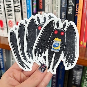 Puede incluir: Una pila de pegatinas en blanco y negro que representan una criatura parecida a una polilla de dibujos animados con ojos rojos y grandes alas. La criatura sostiene un snack Moon Pie. Las pegatinas se sostienen frente a una estantería.