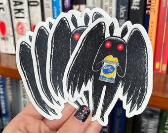 May include: A stack of black and white stickers featuring a cartoon moth-like creature with red eyes and large wings. The creature is holding a Moon Pie snack. The stickers are held in front of a bookshelf.