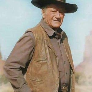 May include: A man wearing a brown leather vest, a brown button-down shirt, and a black cowboy hat. He is also wearing a gold bracelet and a belt with a gold buckle. The background shows a desert landscape.