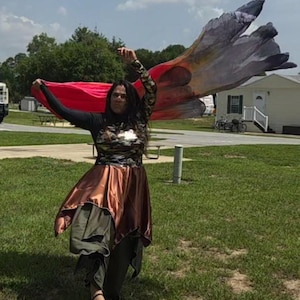 May include: A long-sleeved garment featuring a black bodice with a camouflage-patterned chest panel, paired with a metallic copper-colored skirt and olive green layered trousers. Large, flowing fabric wings in red, orange, and black are attached.
