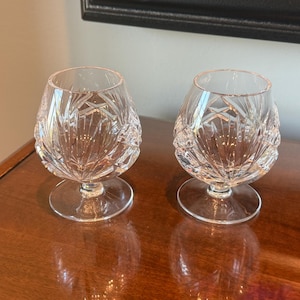 May include: Two clear crystal brandy snifters with intricate cut glass patterns. The glasses have a rounded bowl shape and sit on a short stem with a circular base. The glasses are displayed on a polished wooden surface.