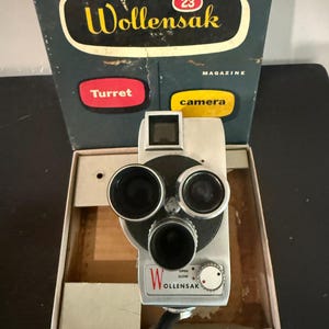 May include: Vintage Wollensak 23 Magazine camera with a silver body and three lenses. The camera is in its original box, which has the word "Wollensak" in a black oval with yellow lettering. The box also has the words "Turret" and "camera".