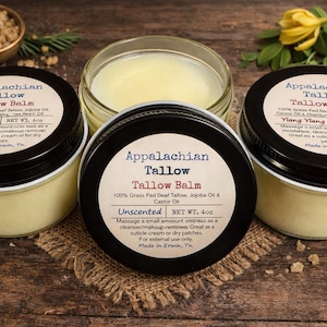 May include: Three jars of Appalachian Tallow Balm with black lids and white labels, arranged on a wooden surface. The labels display the product name, ingredients, and usage instructions. The balm is a light yellow color. The jars are surrounded by greenery and small bowls of grains.