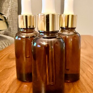 May include: Three amber glass dropper bottles with gold-colored collars and white tops are arranged on a wooden surface. The bottles are cylindrical and appear to be empty. The background is blurred.