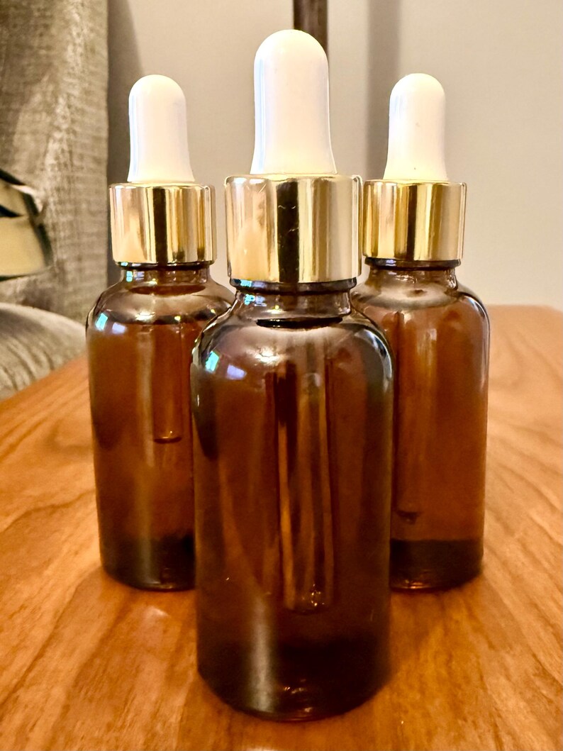 May include: Three amber glass dropper bottles with gold-colored collars and white tops are arranged on a wooden surface. The bottles are cylindrical and appear to be empty. The background is blurred.