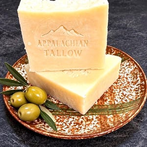 May include: Two rectangular bars of off-white tallow soap stacked on a speckled brown and white ceramic plate. The top bar features an embossed mountain graphic and the text APPALACHIAN TALLOW. Three green olives with leaves sit beside the soap.