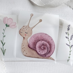 May include: Three separate adhesive decals featuring watercolor illustrations. The central decal shows a light brown snail with a large, dark pink spiral shell. The left decal depicts a single pink flower on a green stem, and the right decal shows a sprig of purple lavender.