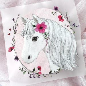 Puede incluir: Una impresión cuadrada con la ilustración de una cabeza de caballo blanco centrada sobre un fondo circular rosa suave. El caballo tiene una crin fluida y una flor rosa brillante detrás de la oreja. Una delicada corona de pequeñas flores rojas y moradas rodea al caballo, acentuada por dos pequeños corazones rojos en el lado izquierdo.