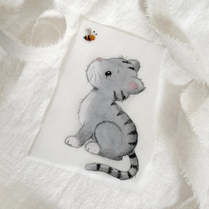 Könnte beinhalten: Ein rechteckiger Druck mit einer Aquarellillustration eines kleinen grauen getigerten Kätzchens mit dunklen Streifen, weißen Pfoten und rosa Ohren, das zu einer winzigen gelb-schwarzen Hummel in der oberen linken Ecke hinaufblickt.