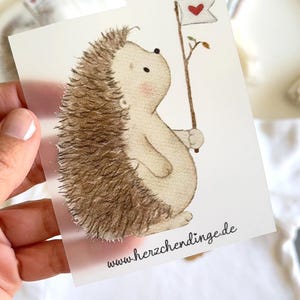 Könnte beinhalten: Eine skurrile Illustration eines Igels, der eine Flagge mit einem Herzen hält. Der Igel hat braune Stacheln und einen hellbeige Körper. Die Flagge ist weiß mit einem roten Herzen. Der Text "www.herzchendinge.de" steht unten.