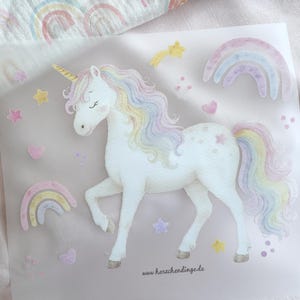 Könnte beinhalten: Eine skurrile Illustration eines weißen Einhorns mit goldenem Horn, fließender pastellfarbener Mähne und Schweif sowie geschlossenen Augen. Das Design umfasst Regenbogen, Sterne und Herzen in sanften Farben. Die Website-Adresse "www.herschendinge.de" ist sichtbar.