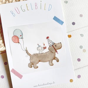 Könnte beinhalten: Eine weiße Karte mit dem Wort "BÜGELBILD" in Pastellfarben. Die Karte zeigt eine Aquarellillustration eines Hundes mit Partyhut, der eine Maus mit einem Cupcake und Luftballons trägt. Die Website-Adresse "www.herzchendinge.de" befindet sich unten.