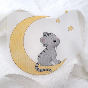 Könnte beinhalten: Eine Aquarellillustration eines kleinen grauen getigerten Kätzchens mit dunklen Streifen, das auf einer großen gelben Mondsichel sitzt. Drei kleine gelbe Sterne befinden sich über dem Kätzchen, das auf einem weißen Hintergrund dargestellt ist.