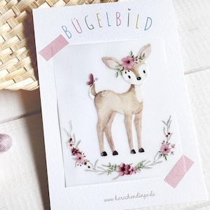 Könnte beinhalten: Aquarell-Illustration eines Rehs mit Blumenkranz und Schmetterling am Schwanz. Das Bild befindet sich auf einer weißen Karte mit dem Wort "BÜGELBILD" in bunten Buchstaben. Ein Blumenkranz umgibt das Reh.