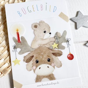 Könnte beinhalten: Ein Bügelbild mit einem skurrilen Weihnachtsdesign. Das Bild zeigt einen Bären und einen Elch, die Geweihe mit Sternen und Ornamenten schmücken. Das deutsche Wort "BÜGELBILD" steht oben. Zwei silberne Sternornamente befinden sich rechts.