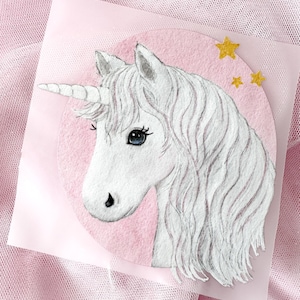 Könnte beinhalten: Eine kreisförmige Illustration eines weißen Einhorns mit spiralförmigem Horn, langer, fließender Mähne und einem blauen Auge vor einem zartrosa Hintergrund. Drei kleine, glitzernde goldene Sterne befinden sich in der oberen rechten Ecke.
