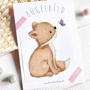 Könnte beinhalten: Aquarell-Illustration eines Fuchswelpen und eines Schmetterlings auf einer weißen Karte. Das Wort "BÜGELBILD" steht oben. Die Karte wird mit rosa Klebeband befestigt. Die Website-Adresse "herzchdinge.de" befindet sich unten.