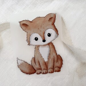 Könnte beinhalten: Ein rechteckiger weißer Aufnäher mit einer Illustration im Aquarellstil, die einen sitzenden Fuchs zeigt. Der Fuchs hat braunes Fell, eine weiße Brust, eine weiße Gesichtsmaske und eine weiß gefärbte Schwanzspitze mit schwarzen Umrandungen.